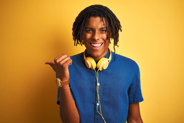 Hombre afroamericano con rastas con auriculares sobre fondo amarillo aislado sonriendo con la cara feliz mirando y apuntando hacia un lado con el pulgar hacia arriba
. - Foto, Imagen
