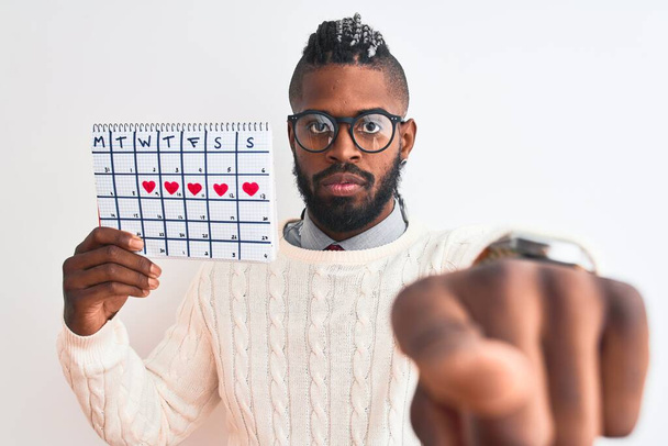 Afro-Amerikaanse man met vlechten houden periode kalender over geïsoleerde witte achtergrond wijzend met de vinger naar de camera en naar u, handteken, positieve en zelfverzekerde gebaar van de voorkant - Foto, afbeelding
