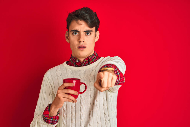 Teenager αγόρι φορώντας πουλόβερ χειμώνα πίνοντας ένα φλιτζάνι καφέ πάνω από απομονωμένο κόκκινο φόντο δείχνοντας με το δάχτυλο στην κάμερα και σε σας, σημάδι στο χέρι, θετική και σίγουρη χειρονομία από το μπροστινό μέρος - Φωτογραφία, εικόνα