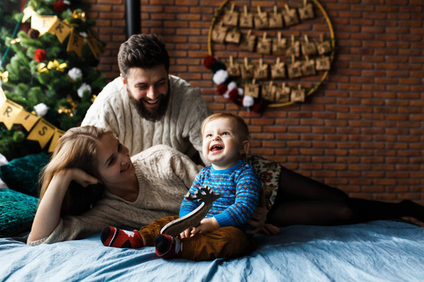 Τα πρώτα Χριστούγεννα. Το χαρούμενο αγοράκι λαμβάνει ένα δώρο από τον Άγιο Βασίλη για πρώτη φορά. Χαρούμενα συναισθήματα γονείς και μωρό. Baby κάθεται στο κρεβάτι, χριστουγεννιάτικο δέντρο, κεριά και διακοσμήσεις στο παρασκήνιο. - Φωτογραφία, εικόνα