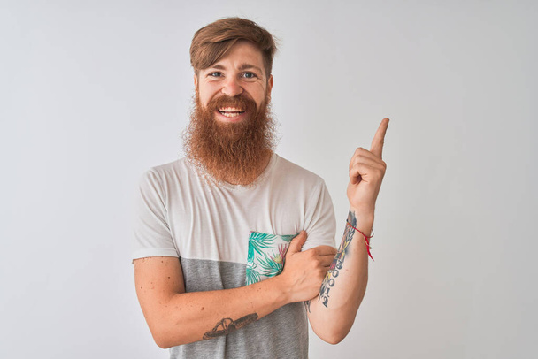 Young redhead irish man wearing t-shirt standing over isolated white background with a big smile on face, pointing with hand and finger to the side looking at the camera. - 写真・画像