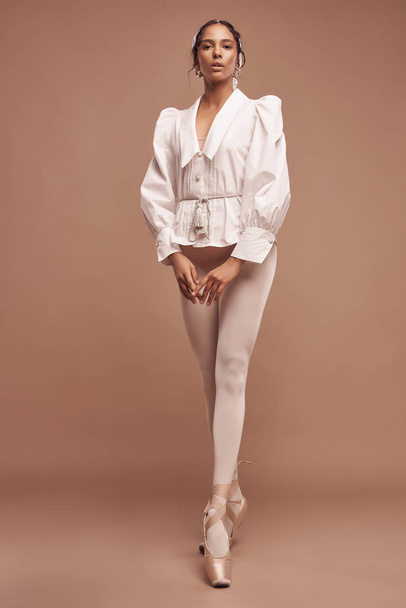 full-length photo of dark-skinned ballerina on brown background wearing white shirt, beige bodysuit, white tights and beige pointes she looks into camera holding hands in front of her - Foto, afbeelding
