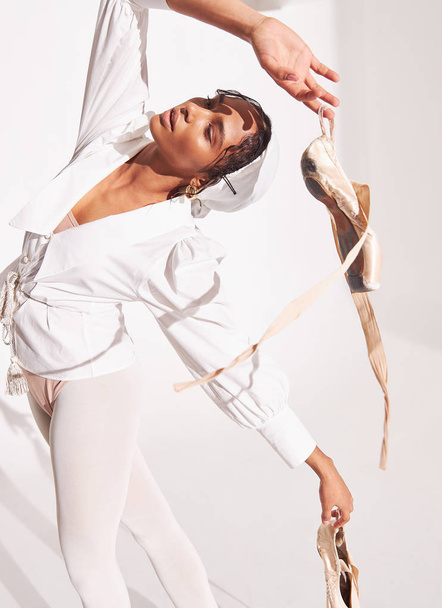 portrait photo of dark-skinned ballerina on white background, she wearing white shirt, beige bodysuit, white tights and holding beige pointes in her arms she leaned back and raised one hand over herself - Fotó, kép