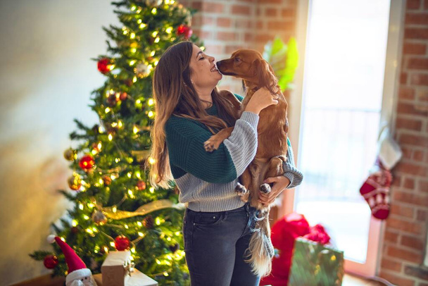Nuori kaunis nainen hymyilee onnellisena ja itsevarmana. Holding ja halaus koira ympäri joulukuusi kotona
 - Valokuva, kuva