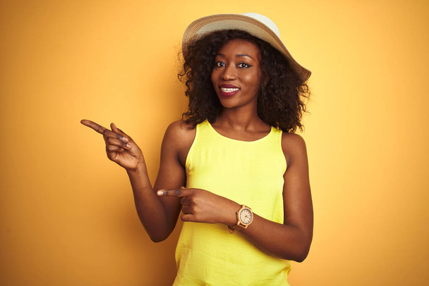 Jeune femme afro-américaine portant t-shirt et chapeau sur fond jaune isolé souriant et regardant la caméra pointant avec deux mains et doigts sur le côté
. - Photo, image