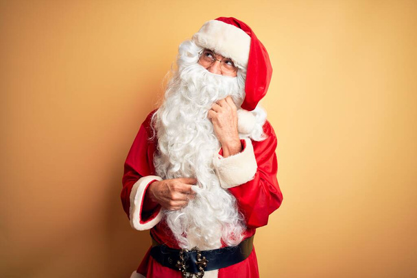 Ein gutaussehender Mann mittleren Alters im Weihnachtsmannkostüm, der vor isoliertem gelben Hintergrund steht, die Hand am Kinn, über Fragen nachdenkend, nachdenklich. Lächeln mit nachdenklichem Gesicht. Zweifelhaftes Konzept. - Foto, Bild