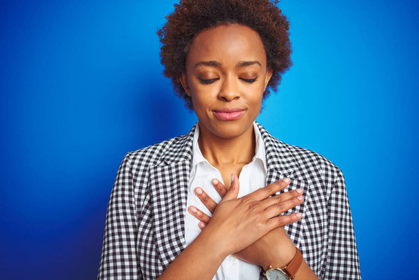 Afro-amerikan yönetici kadın izole mavi arka planda gülümsüyor elleri göğsünde gözleri kapalı ve yüzünde minnet dolu bir jestle. Sağlık kavramı. - Fotoğraf, Görsel