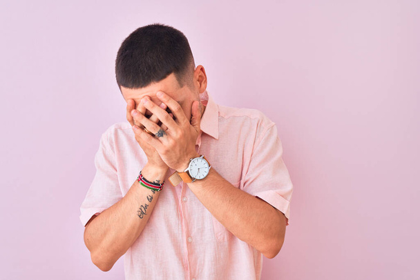 Mladý pohledný muž v růžové košili, stojící nad izolovaným pozadím se smutným výrazem, zakrývajícím tvář rukama, zatímco plakal. Koncept deprese. - Fotografie, Obrázek