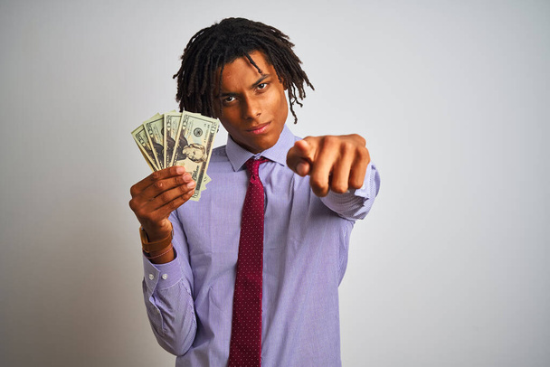 Empresário afro-americano com dreadlocks segurando dólares sobre fundo branco isolado apontando com o dedo para a câmera e para você, sinal de mão, gesto positivo e confiante da frente
 - Foto, Imagem