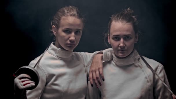 Two young women fencers standing in the dark studio - posing - raising their heads up and looking in the camera - 映像、動画