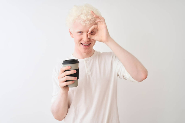 Молодий альбіно блондинка п'є каву, що стоїть над ізольованим білим тлом з щасливим обличчям, посміхаючись, роблячи знак з рукою на око, дивлячись через пальці
 - Фото, зображення