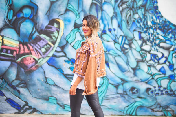 jong mooi meisje dragen bruin jasje staan achteruit over kleurrijke muur in de stad straat - Foto, afbeelding
