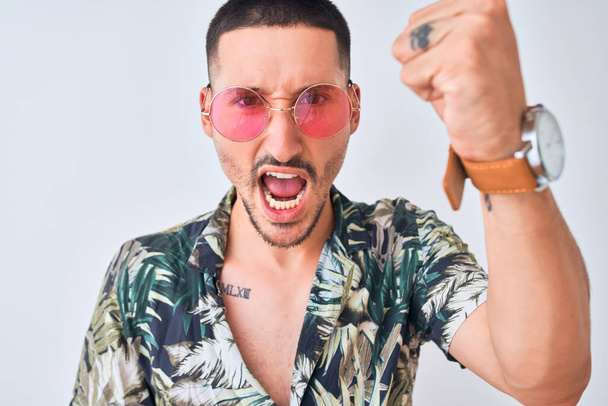 Jonge knappe man draagt Hawaïaanse sumer shirt en roze zonnebril over geïsoleerde achtergrond geïrriteerd en gefrustreerd schreeuwen van woede, gek en schreeuwen met opgeheven hand, woede concept - Foto, afbeelding