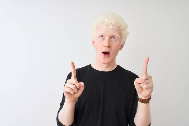 Młody albinos blondyn w czarnej koszulce stojący nad odizolowanym białym tłem zdumiony i zaskoczony patrząc w górę i wskazując palcami i podniesionymi ramionami. - Zdjęcie, obraz