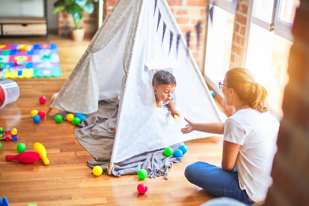 Beautiful teacher and toddler boy sitting on the floor playing inside  tipi at kindergarten - Photo, image
