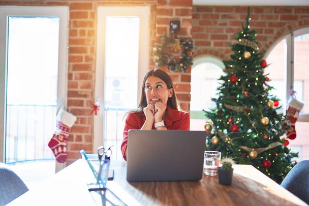 Mooie vrouw zitten aan de tafel werken met laptop thuis rond kerstboom lachen nerveus en opgewonden met handen op kin kijken naar de zijkant - Foto, afbeelding