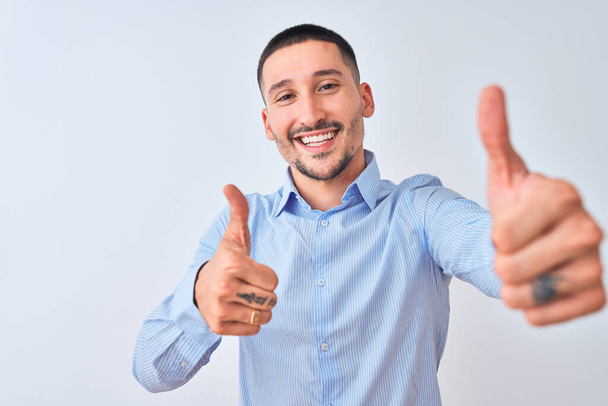 Young handsome business man standing over isolated background approving doing positive gesture with hand, thumbs up smiling and happy for success. Winner gesture. - 写真・画像