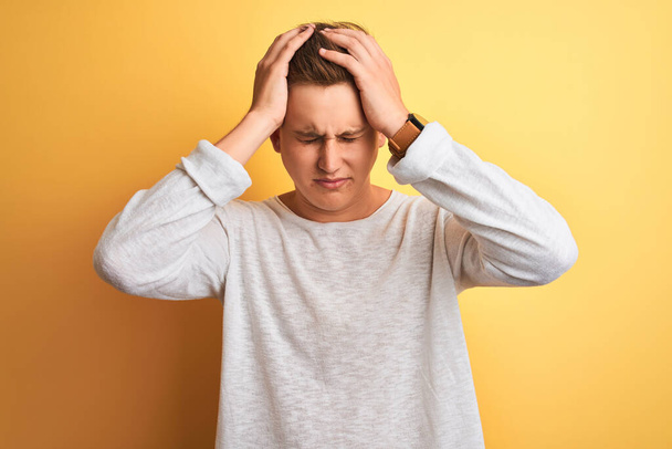 Young handsome man wearing white casual t-shirt standing over isolated yellow background suffering from headache desperate and stressed because pain and migraine. Hands on head. - Фото, зображення