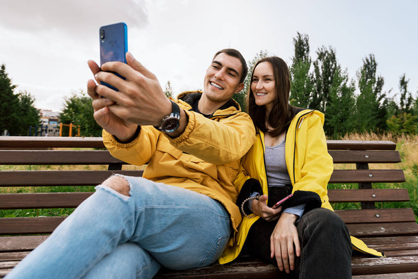 Sarı ceketli, gülümseyen genç bir çift bir selfie çekiyor, akıllı telefon, telefon, sonbahar parkında bir bankta oturuyor. Milenyum kuşakları, cihazdan ayrılmaz. - Fotoğraf, Görsel