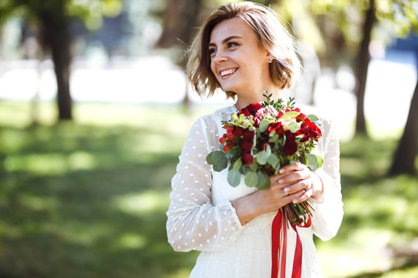 Beautiful bride with a wedding bouquet for a walk in the park. Young bride in the fashionable white dress is incredibly happy. Elegant woman with professional make up and hair style. Wedding day.  - Φωτογραφία, εικόνα