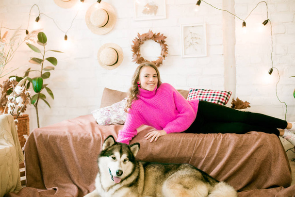 Cute teenage girl with blond curly hair at home, on the bed in the loft with a big malamute dog. People and Dogs. - Foto, Bild