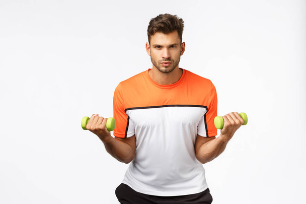 Time get fit. Determined and motivated handsome sexy young male athlete in sportswear, lift dumbbells with assertive focused expression, workout in gym, want strong biceps, white background - 写真・画像