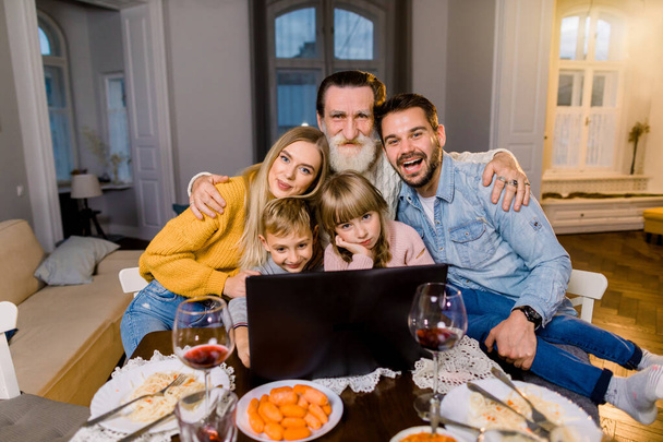 Χαρούμενη οικογένεια γιορτάζει την Πρωτοχρονιά, τα Χριστούγεννα ή την Ημέρα των Ευχαριστιών. Η οικογένεια κοιτάει το λάπτοπ και χαμογελάει. Γιορτινή διάθεση. Νόστιμο φαγητό στο τραπέζι. Ευτυχισμένοι μαζί. - Φωτογραφία, εικόνα
