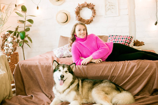 Cute teenage girl with blond curly hair at home, on the bed in the loft with a big malamute dog. People and Dogs. - Photo, image