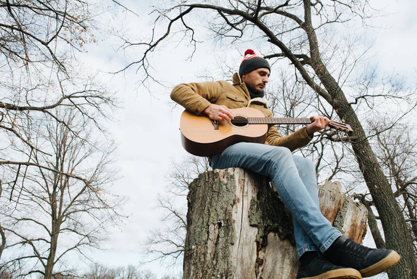 Warmly dressed man sitting on a big stump playing guitar in a co - 写真・画像