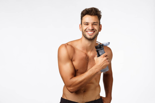 Svalnatý sexy a bezstarostný, šťastný usměvavý sportovec stojící bez trička, držící láhev s vodou a usmívající se potěšený, potěšený po produktivním cvičení, ukazující břišní svaly, balení šesti balení - Fotografie, Obrázek