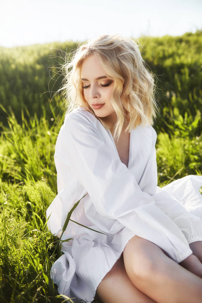 Chica en un vestido blanco largo se sienta en la hierba en un campo. Rubia
  - Foto, imagen