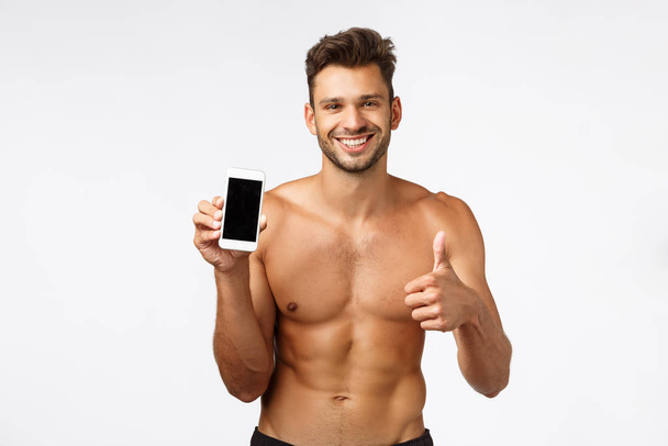 Fešák bez trička koncept dát dobrou radu, doporučujeme stáhnout cvičení fitness tracker app. Sexy sportovec s nahým trupem, perfektní tvar těla, usměvavé potěšení, drží smartphone, show thumbs-up - Fotografie, Obrázek