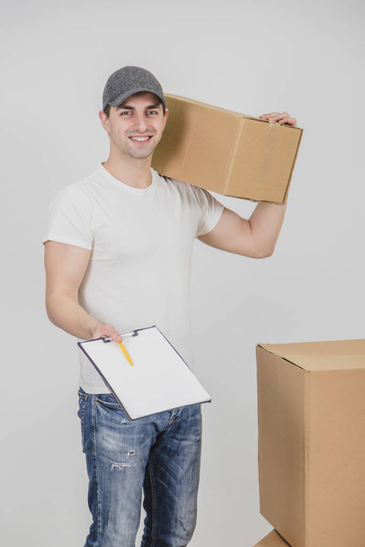 Young delivery man standing with carton box on his shoulder, looking at the camera, smiling, extending a clipboard for signature. - Photo, Image