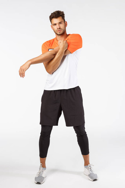 Озил, фитнес и бодибилдинг. Полноразмерный мотивированный красивый спортсмен растягивая руки, разминка перед началом упражнений, расслабление напряжения в мышцах, стоя на белом фоне
 - Фото, изображение