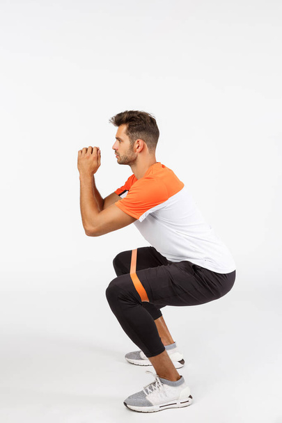 Fitness, health and sport concept. Vertical full-length studio shot active good-looking male athlete, football player in activwear, turn left, perform squats with resistance ropes, workout - Foto, afbeelding
