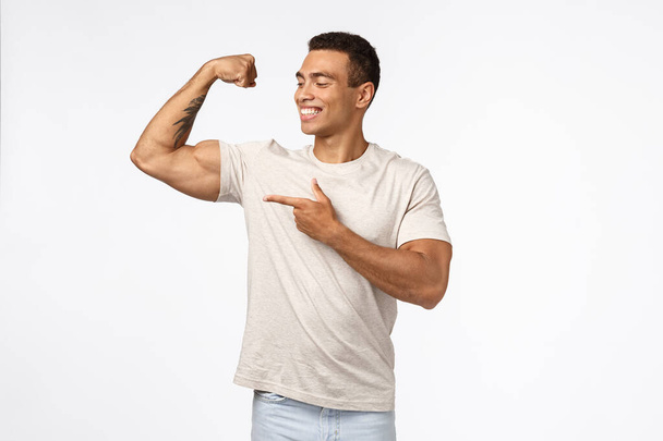 Задоволений, гордий наполегливий молодий бразильський чоловік з сильними м'язами, напружена піднята рука, усміхнені і вказуючі біцепси гордо, в захваті від власного ідеального тіла, тренування в спортзалі, показуючи результати спорту
 - Фото, зображення