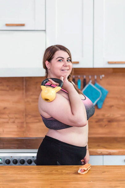 sport with unhealthy food. combination of active life with fast food. close up photo. fat woman showing kettlebell and hot dog - Fotó, kép