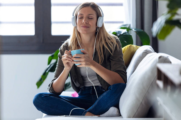 Pretty young woman listening to music with headphones while drinking cup of coffee on sofa at home. - Photo, Image