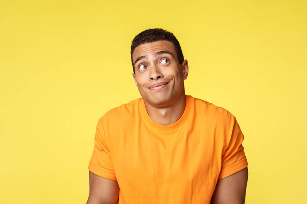 Unsure, careless and unbothered handsome brazilian guy in casual t-shirt, shrugging indecisive, look up and smirk clueless, dont know, trying avoid eye contact as hiding something, yellow background - Photo, image