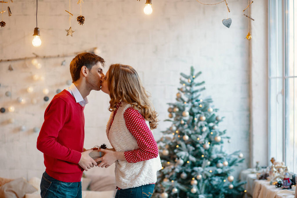 Young couple kissing while holding together a present box, with Christmas decorations in background - Photo, Image
