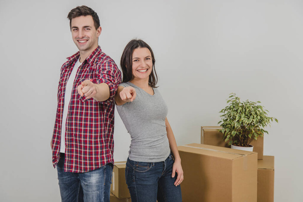 Happy man and woman standing side by side near stack of carton boxes in their new apartment, pointing their fingers at the camera, smiling. - Fotoğraf, Görsel