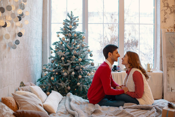 Happy loving couple enjoying Christmas. Young man kissing his girlfriend on forehead touching her hands sitting on a bed with decorated tree and window on background - Photo, Image