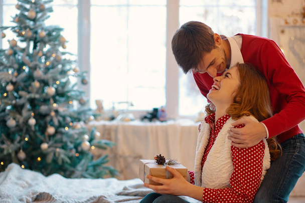 Joyeux couple amoureux profitant de Noël. Jeune homme touchant son nez de petite amie avec son nez tenant un cadeau avec fenêtre sur fond
 - Photo, image