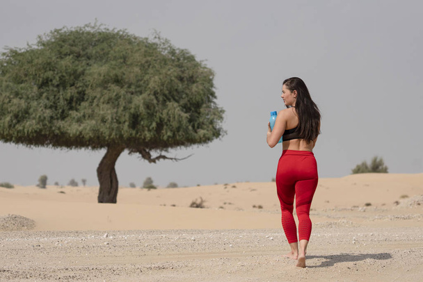 Krásná brunetka fitness model na sobě jasně červené punčocháče a černé sportovní podprsenka chůze směrem ke stromu v horké poušti oblasti na jasný slunečný den drží láhev modré vody - Fotografie, Obrázek