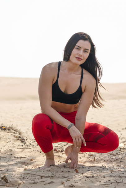 Beautiful brunette fitness model wearing bright red tights and black sports bra  in a squat pose in  a hot Arabian desert on a bright sunny day - Photo, Image