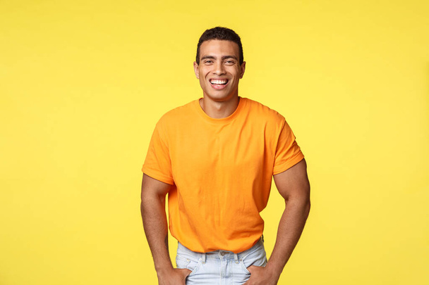 Komea iloinen hymyilevä, maskuliininen mies oranssissa t-paidassa nauraa iloisesti, hymyilee ystävällisellä positiivisella ilmeellä, pitää kädet taskuissa rennosti seisoen uudella vuodella, keltainen tausta
 - Valokuva, kuva