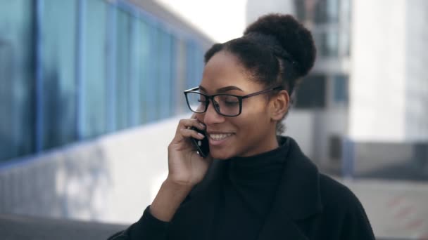 Cheerful businesswoman in glasses talking by smartphone. Emotional young African American businesswoman talking by cell phone being outside on the street. Connection concept - Кадри, відео