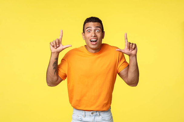 Fascinado, emocionado joven caucásico hombre en camiseta naranja, cámara de mirada asombrado e impresionado, apuntando hacia arriba como ver oferta increíble promo, de pie fondo amarillo sorprendido
. - Foto, imagen
