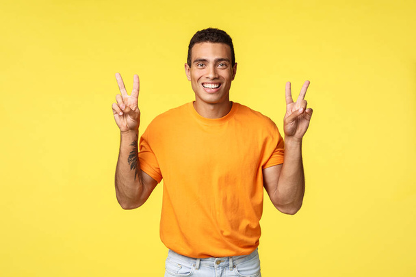 Handsome joyful young male in orange t-shirt, smiling upbeat, make peace gesture or quotes sign, standing yellow background, express positivity and optimism, standing yellow background - Photo, Image