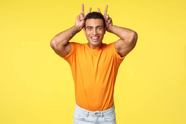 Roztomilý mužský a hloupý hispánský přítel v oranžovém tričku, udělat mírové gesto, králičí uši za hlavou, radostně se usmívající, napodobit králíka, aby si hrál s neteří přes žluté pozadí - Fotografie, Obrázek